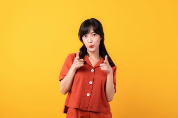 Portrait beautiful young asian woman happy smile dressed in orange clothes showing pointing finger hands gesture to camera isolated on yellow studio background. Attention to nice product