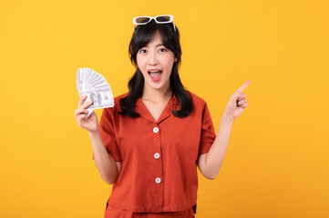 Portrait young beautiful asian woman happy smile dressed in orange clothes holding cash money and celebrate her success isolated on yellow background. Be rich concept.