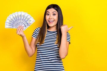 Photo of young woman wear striped dress hold dollars banknotes direct finger empty space invest company isolated on yellow color background