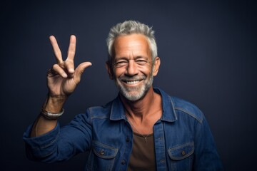 Fototapeta premium Lifestyle portrait photography of a satisfied mature man making a peace and love gesture with the fingers against a navy blue background. With generative AI technology