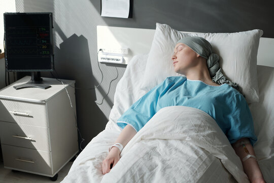 Young Woman In Headscarf Sleeping Under Blanket In Hospital Bed Next To Black Monitor Of Computer While Having Chemotherapy Procedure