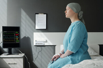 Young recovering female patient of oncological clinics in headscarf and blue uniform sitting on bed...