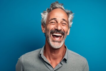 Obraz premium Close-up portrait photography of a grinning mature man placing the hand over the mouth in a laughter gesture against a soft blue background. With generative AI technology