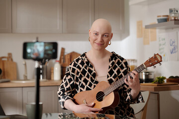 Young smiling woman with bald head playing guitar and looking at smartphone camera while recording new music lesson for online audience