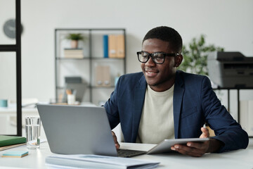 Fototapeta na wymiar Young successful African American businessman in eyeglasses and elegant suit using mobile gadgets while sitting by workplace in front of laptop