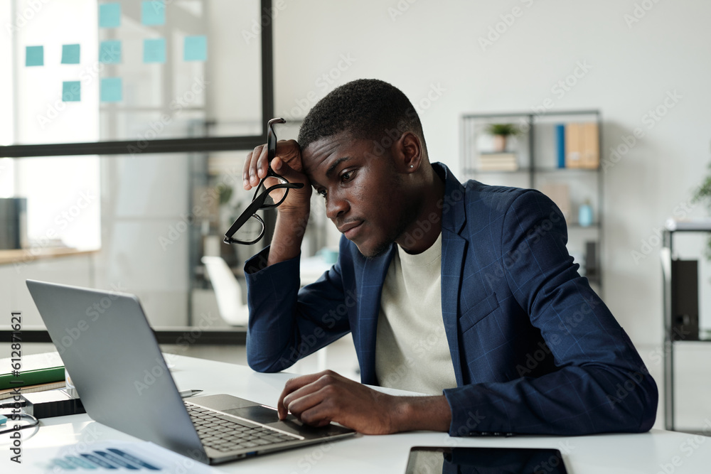 Poster young tense african american businessman sitting by workplace in front of laptop in office and looki