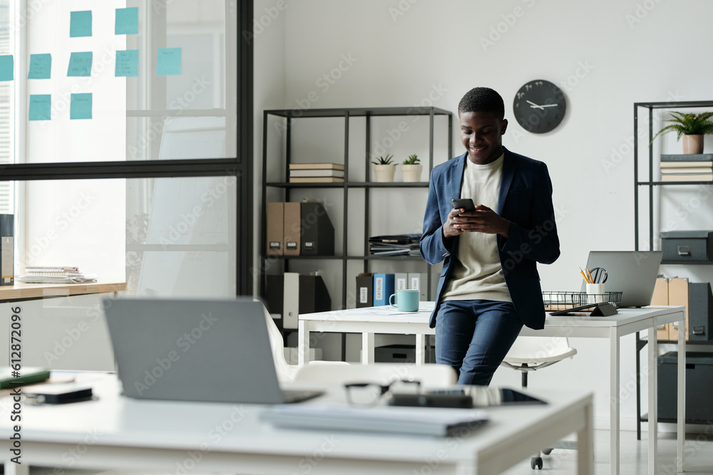 Aufkleber Young smiling African American businessman in formalwear using mobile phone while texting in front of camera in openspace office