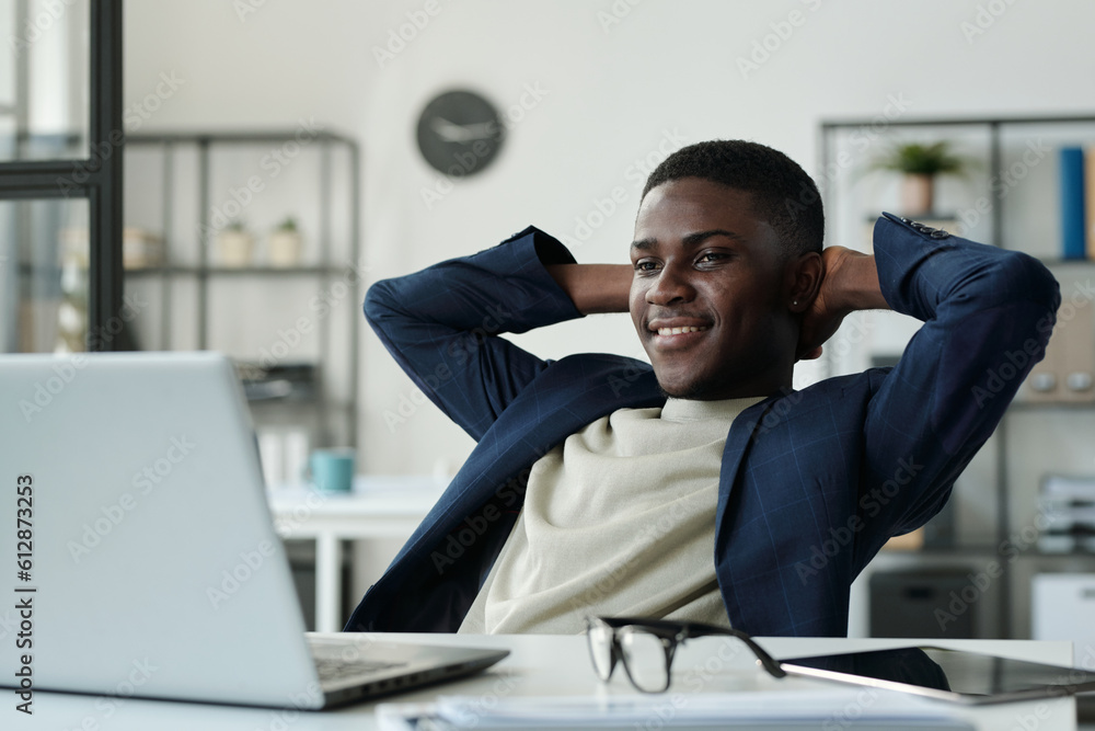 Poster young restful businessman keeping hands behind head and looking at laptop screen while watching onli