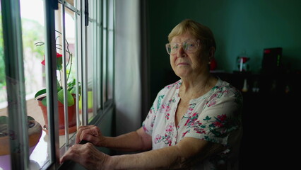 Portrait of an elderly woman looking at camera standing by window at home. Authentic domestic old age lifestyle of senior person in 80s