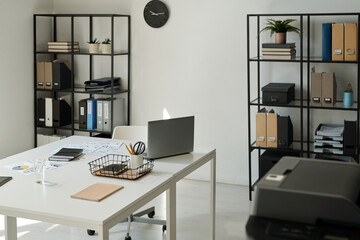 Part of spacious openspace office with laptop, basket with pencils, notebooks and other supplies on workplace of white collar worker