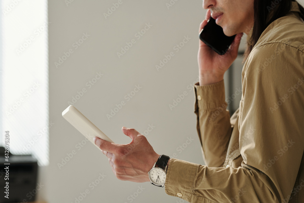 Poster Side view of young director of business company with mobile phone by ear and tablet scrolling through online data and talking to subordinate