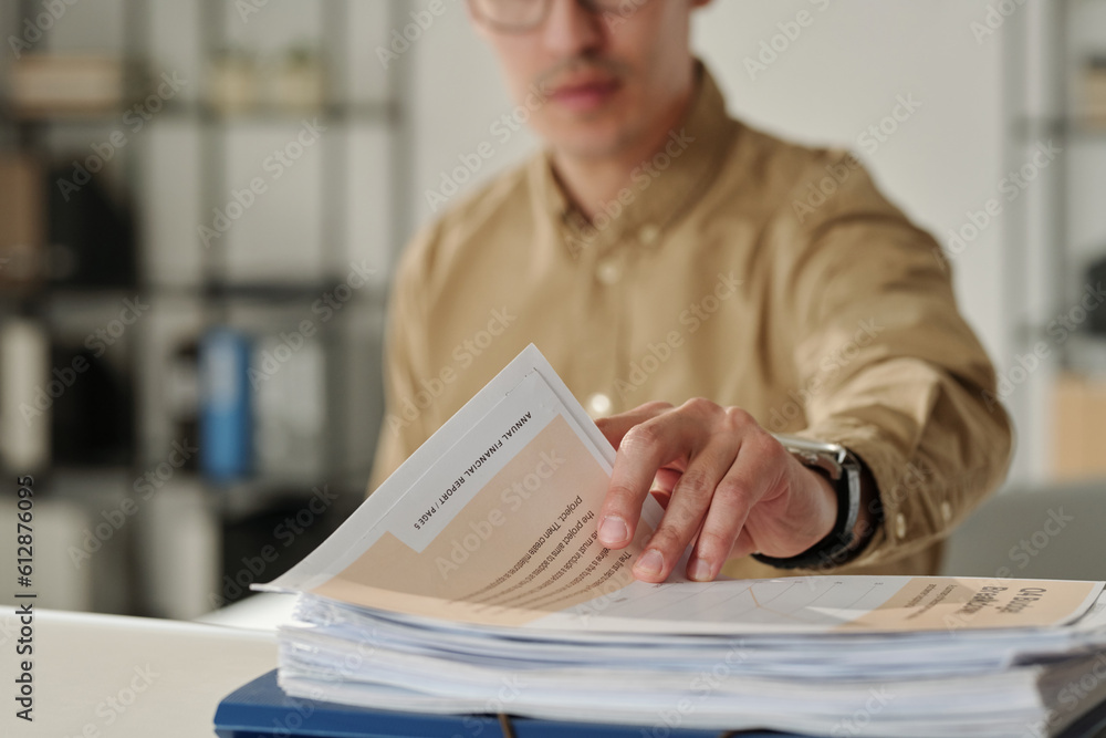 Poster Hand of young male economist or auditor taking financial document from stack of papers while analyzing data during work over new project
