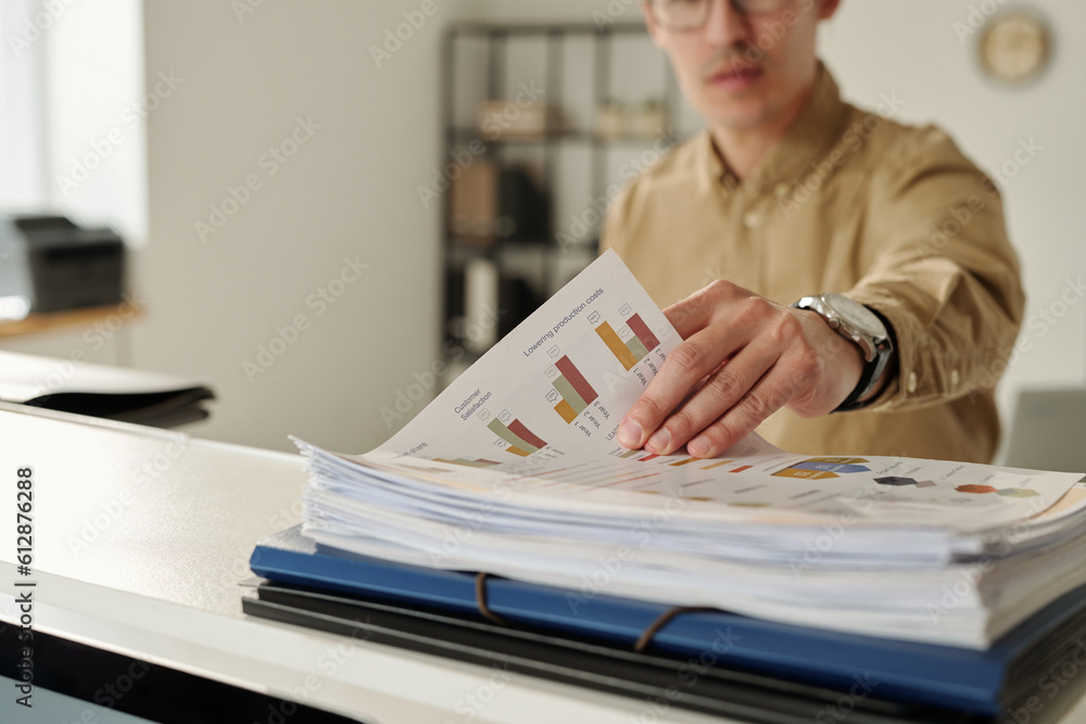 Aufkleber Hand of young analyst or marketing manager taking document with financial charts from stack of papers before analyzing graphic data