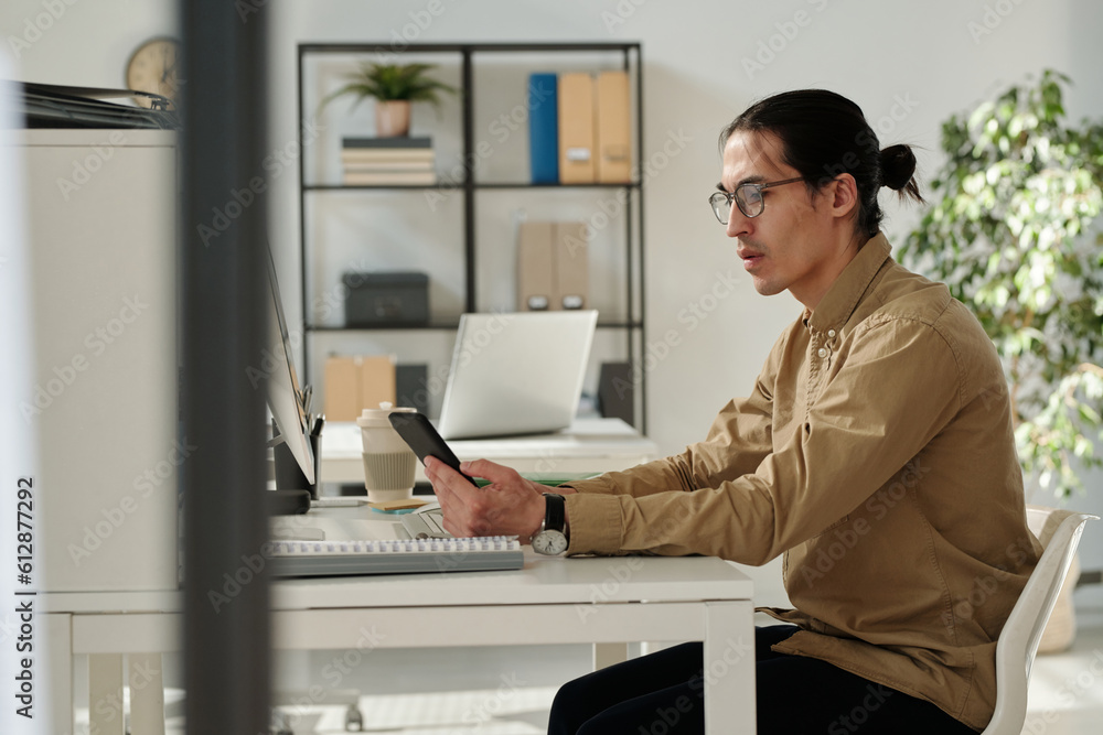 Aufkleber Side view of young serious analyst or broker texting in mobile phone while sitting by workplace in front of computer and working over new project