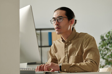 Young Asian male entrepreneur looking at online financial data on computer screen while analyzing it and preparing report for seminar