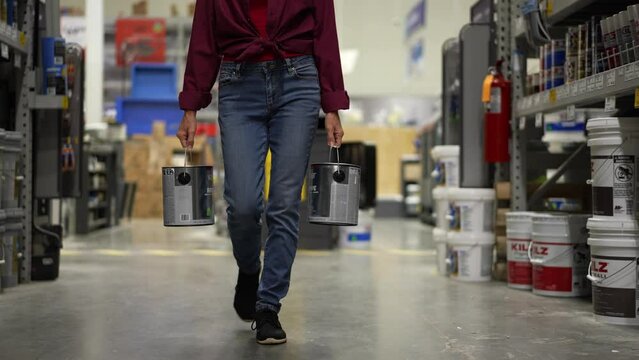 Slow motion of woman shown from waist down, carrying to cans of paint walking down paint aisle in hardware store