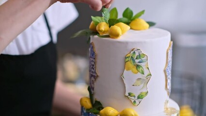 Focused blonde woman putting confit yellow lemons on large wedding cake standing at table in modern kitchen preparation process of homemade custom confectionery from lactose free products