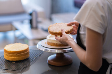 Confectioner in black apron connecting organic cake layers with fresh white cream standing at table in modern kitchen detailed process of preparing delicious pastry closeup