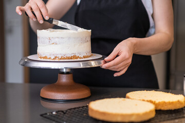 Food blogger confectioner preparing delicious lactose free cake for event at home woman in black apron standing at kitchen table demonstrates smearing white cream on handmade confectionery