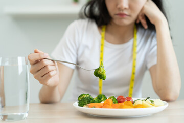 unhappy asian women is on dieting time looking at broccoli on the fork. girl do not want to eat vegetables and dislike taste of broccoli.