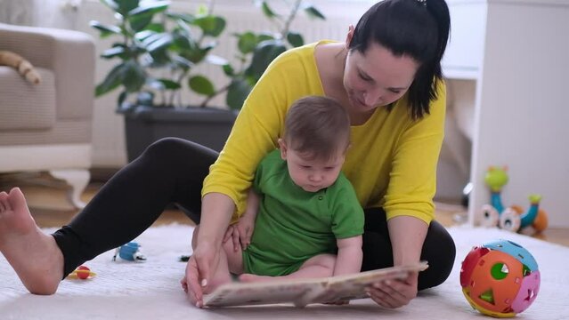 Caring mother showing focused little son book with colorful pictures of living creatures sitting on soft rug in nursery woman educating baby boy at home