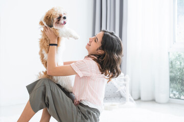 Caucasian teenage girl playing with shih tzu puppy dog at home. Young beautiful woman sitting on floor, smiling, having fun holding little fluffy dog pet with love and care