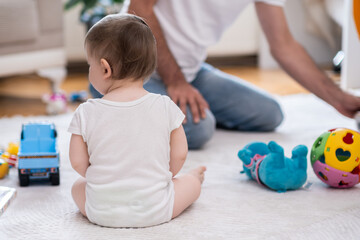 Adorable little boy in white bodysuit sitting on fluffy rug near daddy collecting scattered toys on floor sweet baby enjoying spending time with father and resting at home