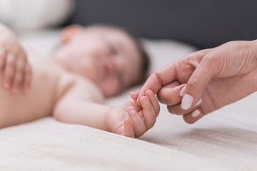 Sleepy little boy grabbing finger of mother with tiny hand lying on soft bed with white sheet woman expressing love and affection to son in bedroom closeup