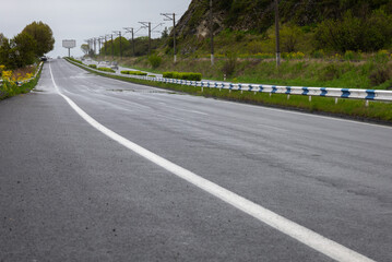 On a beautiful rainy spring day, the highway with puddles and wet asphalt runs alongside picturesque hills covered with lush green grass