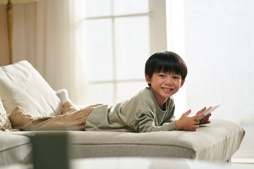 happy cute little asian boy lying on front on family couch with digital tablet in hands