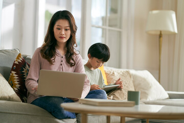 young asian mother working on laptop while son playing computer game using digital tablet at home - obrazy, fototapety, plakaty