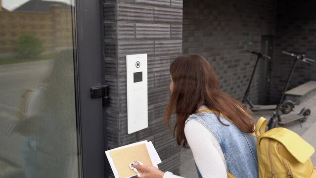 Young brunette woman with keys, letters and backpack dials the code for a call over the intercom. Teenager schoolgirl waving and looking at the camera of the electronic lock. Girl at the lock door