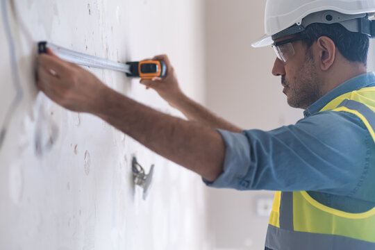 Focused engineer using tape-line to measure distance on wall for socket hole man in helmet and vest working on renovation in living room construction tools and uniform