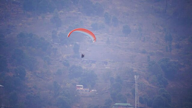 zoomed shot showing para gliding people with a bright orange white glider in the middle of hills mountains near nanital bir billing manali showing a popular adventure sport for tourists