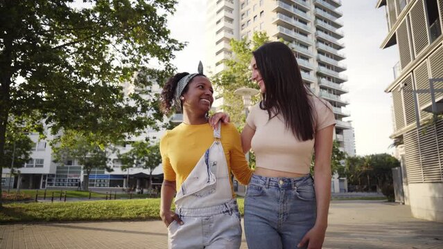Multiracial cute lesbian couple in love walk embracing laughing on city street. Two cheerful young friends stroll hugging on summer day in park. Gay women and relationships in youth generation z.