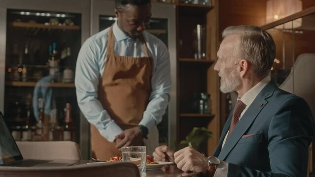 Waist up of mature Caucasian businessman in elegant suit having business lunch in modern restaurant. Young African American waiter serving main dish to him putting plate on table
