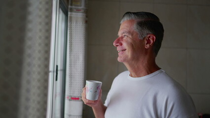 Contemplative middle-aged man standing by kitchen window sipping coffee during morning ritual, domestic everyday lifestyle of gray hair man sips warm drink