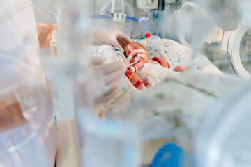 Unrecognizable hand in gloves of nurse or doctor taking care of premature baby placed in a medical incubator. Neonatal intensive care unit in hospital.