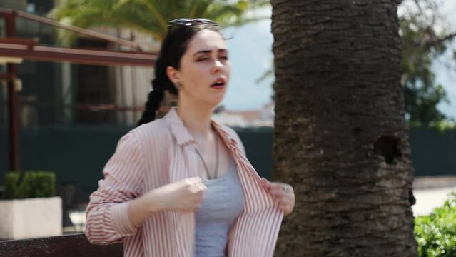 Young Caucasian suffering woman is sitting on bench fanning herself with shirt because of high hot temperature. Summer season. Concept of overheating and sunstroke