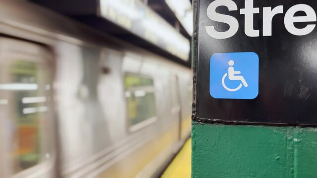 A closeup view of a Manhattan subway sign with a handicap icon as a train approaches the station.  	