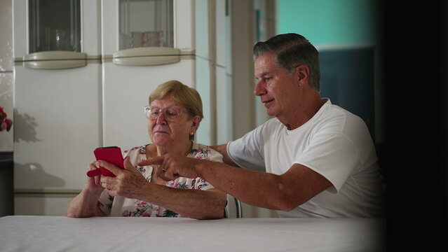 Candid Middle-Aged son helping elderly mother with modern technology in home kitchen. Authentic real people of different generations interacting with phone screen