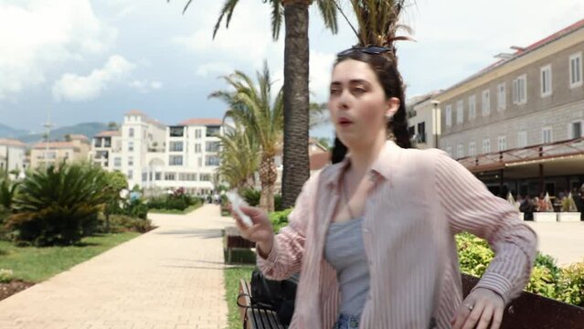Portrait of young Caucasian suffering woman is sitting on bench wipes sweat on face of high hot temperature. Summer season. Concept of overheating, thirst and sunstroke