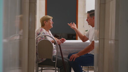 Candid domestic scene of middle-aged son in conversation with elderly mother at home. Authentic...