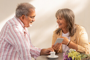 Cheerful european mature couple sit in cafe with flowers drinking coffee, woman show smartphone