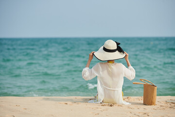 Fototapeta na wymiar Young woman sitting on sandy beach in satin shirt and hat and looking at seascape