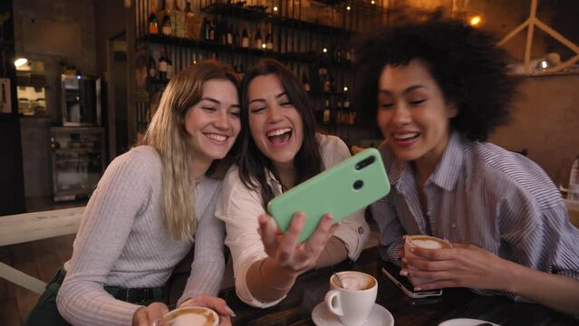 Three friends try out different photo filters with cell phone having fun drink at coffee shop. Smiling happy girls looking at mobile making faces together. Young millennial women and social networks.