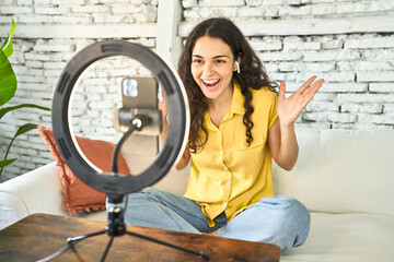 A young Caucasian influencer, creatively recording a vlog on her sofa with a ring light and mobile,...