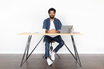 Happy young indian guy sitting at home office, using laptop