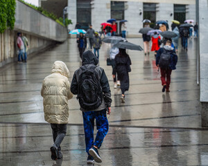Authentic young man and girl with hoods without umbrellas walk in the rain along the city street against the background of a crowd of citizens with umbrellas, soaked passers-by on the city street