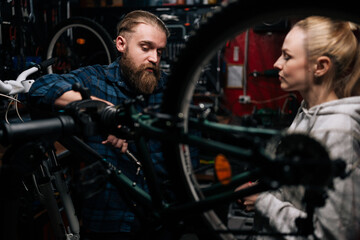 Obraz na płótnie Canvas Serious cycling repairman with beard communicating with attractive blonde female client, talking problem of bicycle, detected during diagnostics in repair shop with dark interior, standing behind MTB.